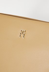 Väri ruskea nahkalaukku, jossa on tekstuuripinta ja kultasävyinen logokoriste. Muotoilu on virtaviivainen ja minimalistinen.