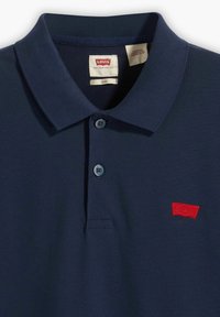 Navy blauwe polo met een drieknoopsluiting, geribbelde kraag en een rood geborduurd logo op de linkerkant van de borst. Textuurdesign van de stof.