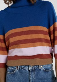 Sininen ja raidallinen neulepaita, jossa vaakasuoria raitoja ruskeassa, vaaleanpunaisessa ja beigen sävyissä, korkealla kauluksella ja resorimaisella pinnalla. Yhdistetty sinisiin farkkuihin.