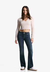 Light pink, ribbed knit crop sweater with long sleeves and button detail. Dark blue flared jeans with a high waist. White sneakers.