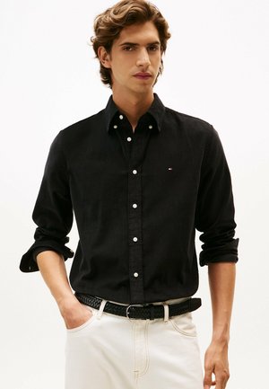 Black button-up shirt with long sleeves, featuring a textured fabric, white buttons, and a small logo on the chest. Worn with a braided black belt.