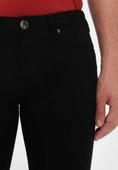 Schwarze Jeans aus elastischem Stoff, mit schmaler Passform, Gesäßtaschen und einem Metallknopfverschluss. Sichtbare Nähte betonen das Design.