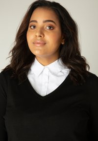 Zwarte V-hals trui gelaagd over een witte blouse met kraag; zachte stoftextuur, aansluitend ontwerp en zichtbare knoopdetails op de blouse.