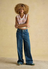 Ärmelloses Strickoberteil in Creme mit mehrfarbigen geometrischen Mustern. Kombiniert mit hochgeschnittenen, weiten blauen Jeans und grünen Sneakers.