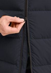 Marine blauwe pufferjas met een zwarte rits. Hand die de rits trekt, met zichtbare gequilte textuur en warm, isolerend materiaal.