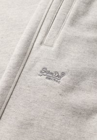 Tissu gris clair avec logo SuperDry® cousu et un détail de couture verticale à proximité.