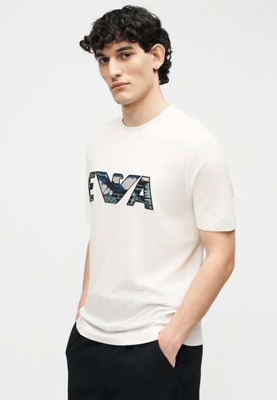 Νέος άνδρας με κατσαρά μαλλιά φοράει λευκό πουκάμισο με κοντά μανίκια με μεγάλα γράμματα μοτίβου "FIVA" και μαύρο παντελόνι, με τα χέρια στις τσέπες.