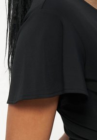 Gros plan sur le haut du bras et l'épaule d'une personne portant un haut noir à manches courtes avec des détails de coutures visibles.