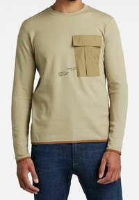 Beige Langarmshirt aus strukturiertem Stoff, mit einer vorderen Cargo-Tasche und bedruckten Details in Schwarz, sowie braunen Akzenten am Saum und an den Bündchen.