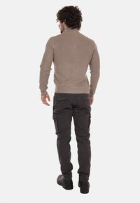 Maglione beige lavorato a maglia con colletto alto, abbinato a pantaloni cargo scuri con tasche laterali e una trama liscia. Stivali completano il look.