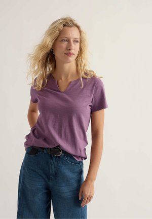 Blonde woman with wavy hair wearing a purple short-sleeve V-neck shirt, dark blue jeans, and a black belt, standing against a plain background.