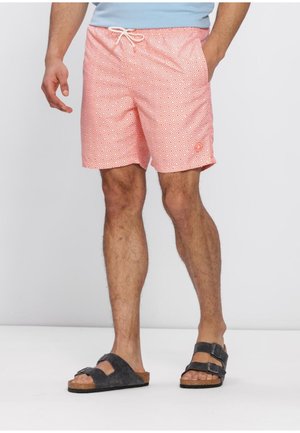 Man die koraalgekleurde zwembroek met patroon en zwarte sandalen met dubbele bandjes draagt, staand tegen een effen achtergrond.