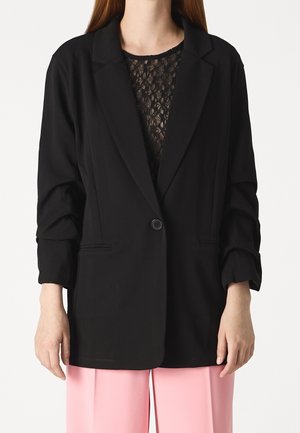 Vrouw draagt een zwart colbert met één knoop over een zwart kanten top met lange mouwen en roze broek, afgebeeld van nek tot dijen.