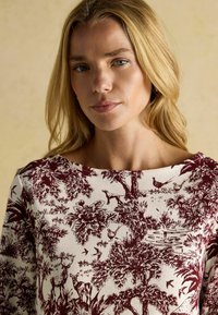 Top s marhuľovým a krémovým vzorom so botanickým dizajnom a zvieratami, rebrovanou textúrou a širokým výstrihom, zobrazený na svetlom pozadí.