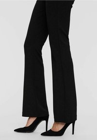 Pantalon noir évasé à la texture lisse, associé à des escarpins noirs en daim pointus avec des talons aiguilles et un design élégant.
