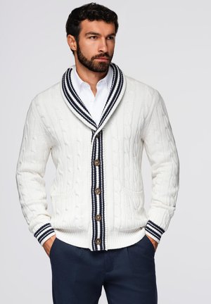 White knitted cardigan with navy blue striped shawl collar, large buttons, and two front pockets. Textured cable pattern throughout.