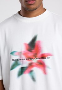 Camiseta blanca de algodón con un gráfico floral difuminado en rojo y verde, y el texto "El Jardín Botánico, Central Park, NY" impreso debajo.