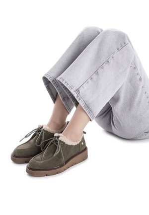Zapatos de ante verde con un forro de felpa beige y cordones a juego, combinados con pantalones de pierna ancha gris claro, en posición sentada.