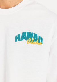 Fehér pamut póló, ívelt türkiz "HAWAII" nyomattal és alatta sárga "Aloha" kiegészítéssel. Sima tapintású, rövid ujjú.