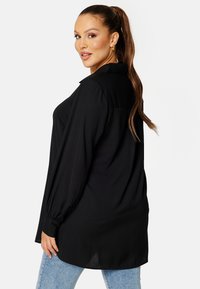 Blusa nera con colletto e maniche lunghe arricciate. Tessuto liscio, vestibilità ampia e orlo posteriore più lungo. Abbinata a jeans blu.