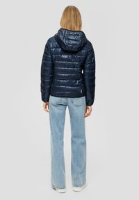Granatowa pikowana kurtka puchowa z kapturem, z pikowanym wzorem, błyszczącą fakturą i lekko krótkim krojem, zestawiona z jasnoniebieskimi dżinsami.