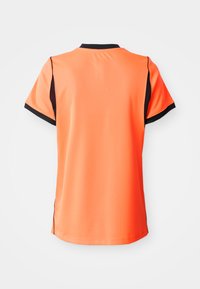 Spilgti oranža sporta krekls ar melnu apdari apkaklē, piedurkņu malās un sānu paneļos, redzams no aizmugures uz vienkārša gaiša fona.