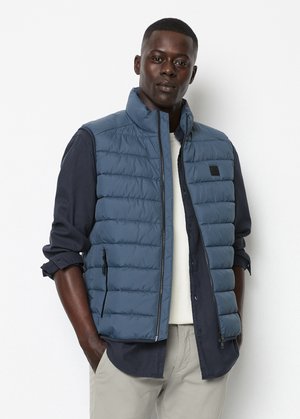 Gevoerde blauwe vest met hoge kraag, ritssluiting aan de voorkant en twee zijzakken, over een donker shirt en lichte broek. Gelaagd, gewatteerd ontwerp.