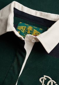 Maillot de rugby vert foncé et bleu marine avec col blanc, bouton unique et étiquette Superdry jaune à l'intérieur du col.