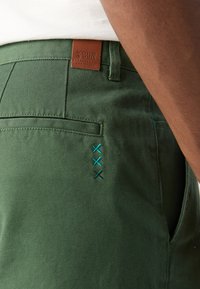 Pantaloni in cotone verde con un dettaglio di ricamo blu decorativo e un'etichetta in pelle con "SCOTCH & SODA" sulla vita.