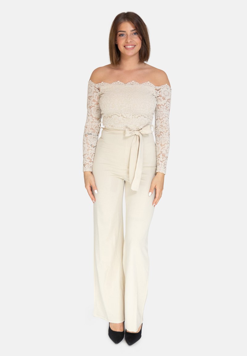Spitzenoberteil mit schulterfreien Ärmeln in hellem Beige und langen Ärmeln. Hochgeschnittene, weite Creme-Hosen mit einer gebundenen Schleife an der Taille. Schwarze High Heels.