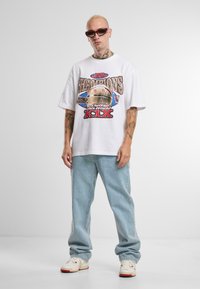 Hvit grafisk t-skjorte med "Champions" trykk, kombinert med lyseblå baggy jeans. Tilbehør med røde solbriller og hvite sneakers.