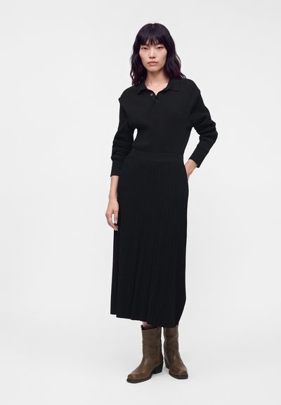 Femme aux longs cheveux noirs portant une robe noire à manches longues et col, ainsi que des bottes marron mi-mollet, debout devant un fond clair uni.