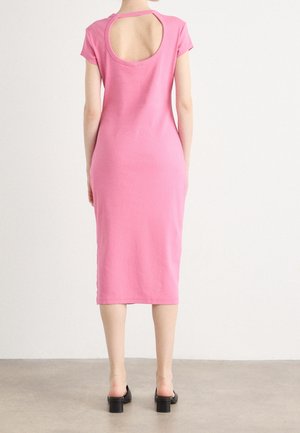 Femme portant une robe midi rose à manches courtes avec une grande découpe ronde dans le haut du dos, associée à des chaussures à talons noires.