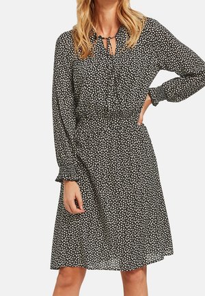 Femme portant une robe noire longueur genou avec des motifs de feuilles blanches éparpillées, des manches longues, des poignets élastiques et une petite encolure goutte d'eau avec un détail de lien.