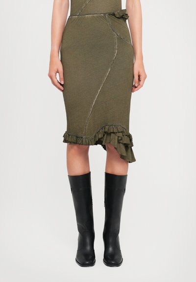 Robe ajustée vert olive avec un ourlet asymétrique, un bas volanté et des détails cousus. Associée à des bottes en cuir noir montantes.