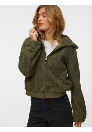 Donna con capelli castano chiaro che indossa una giacca in pile verde oliva con zip e pantaloni neri, tenendo con una mano il colletto della giacca.