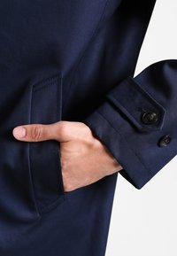 Ręka umieszczona w przedniej kieszeni granatowego płaszcza z długim rękawem, z detalami guzików na mankietach.