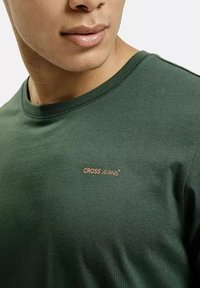Ciemnozielona bawełniana koszulka z okrągłym dekoltem i małym pomarańczowym logo "CROSS JEANS" na piersi. Gładka tekstura, codzienny design.