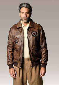 Veste bomber en cuir brun avec des badges, fermeture éclair et deux poches avant. Poignets et col côtelés ; portée sur un pull beige et un pantalon beige.