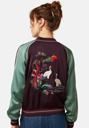 Vrouw met bruin haar in een knot draagt een satijnen bomberjack met mintgroene mouwen en geborduurde kraanvogels, een boom en bloemen op de achterkant.