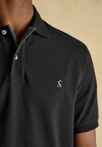 Homme portant un polo noir à manches courtes avec deux boutons et un petit logo animalier brodé en blanc sur le côté gauche de la poitrine.