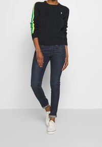 Svart kabelstickad tröja med gröna och gula ränder på ärmarna, matchad med mörkblå skinny jeans och vita sneakers.