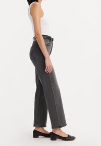 Kvinna i vit ärmlös topp, högmidjade svarta jeans med blekt utseende och svart bälte, samt svarta lågklackade ballerina-skor, stående i profil.