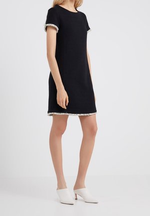 Kobieta w krótkiej, czarnej sukience typu shift z białą frędzlą na rękawach i dole sukienki, zestawiona z białymi mule na obcasie.