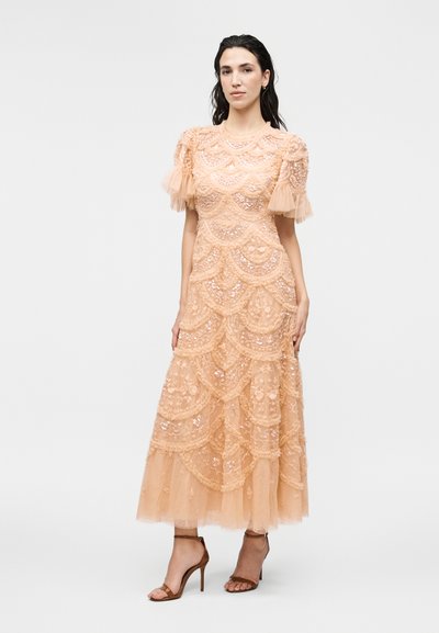 Γυναίκα με σκουρόχρωμα μαλλιά φοράει φόρεμα μέσης μήκους σε ροδακινί δαντέλα με φουσκωτά μανίκια και βολάν στο τελείωμα, συνδυασμένο με καφέ σανδάλια με τακούνι, στέκεται μπροστά σε λευκό φόντο.