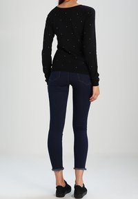 Suéter negro de manga larga con piedras brillantes dispersas, combinado con jeans skinny azul oscuro con dobladillo deshilachado y zapatos negros.