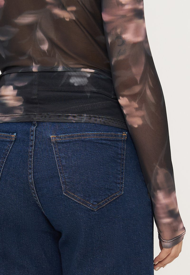 Durchsichtige schwarze Bluse mit floralen Mustern und langen Ärmeln, kombiniert mit dunkelblauen Jeans aus Denim, die über Gesäßtaschen und dezente Nähdetails verfügen.