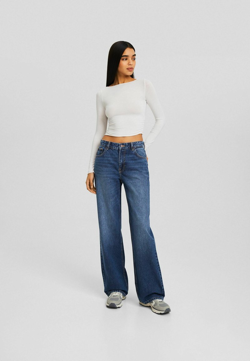 Weißes langärmliges Cropped-Top kombiniert mit weiten blauen Jeans und grauen Sneakers. Einfaches Design, enganliegendes Oberteil und lockere Jeans.