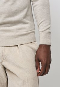 Beiger Pullover mit gerippten Bündchen und Saum, kombiniert mit beigen Cordhosen mit vertikalen Streifen. Hand seitlich positioniert.