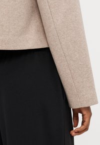 Beige filtjacka med rak skärning och långa ärmar, kombinerad med ett svart, slätt material under. Närbild på ärm och hand.